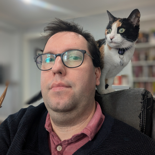 Ryan in an office chair with his calico cat Ciri on the chair behind his head.